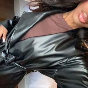 Svart lång kappa i syntetmaterial - Snygg svart lång kappa i syntetmaterial med knytbälte i midjan. Perfekt för en stilren look. Passar bra till både vardag och speciella tillfällen.