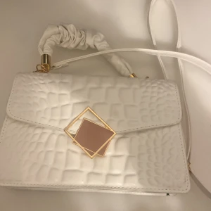 Vit axelväska med geometrisk detalj - Snygg vit axelväska med quiltad textur och unik geometrisk detalj i guld och beige på framsidan. Väskan har en justerbar axelrem med en dekorativ rynkad del. Perfekt för att ge en stilren touch till din outfit. Perfekt för sommaren! 