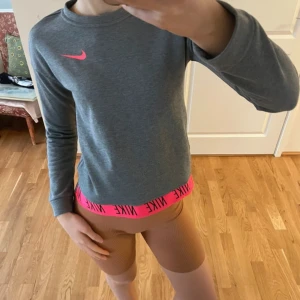 Grå tröja från Nike med rosa detaljer - Säljer en grå tröja från Nike med rosa logga och elastisk kant med Nike-text. Tröjan har långa ärmar och en sportig look. Perfekt för en aktiv livsstil. Passar en XS