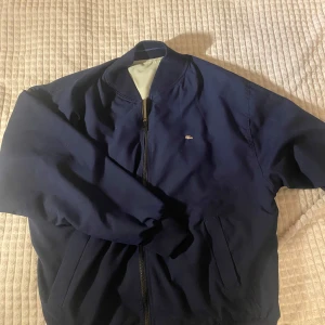 Reversible Lacoste jacka - Snygg vändbar bomberjacka från Lacoste i marinblått och beige. Jackan har en dragkedja framtill och ribbade muddar vid ärmslut och nederkant. Perfekt för att variera din stil med två färger i en jacka. Den är väldigt skön och perfekt för våren. Pris kan diskuteras, byten kan vara intressant 