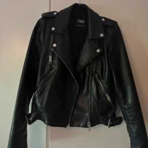 Svart skinnjacka från Zara - Snygg svart skinnjacka från Zara med dragkedjor och knappar i silver. Jackan har en klassisk bikerstil med bälte i nederkant och långa ärmar. Perfekt för en cool look!