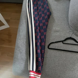 Mörkblå mjukisbyxor med GG-mönster från Gucci - Snygga mörkblå byxor från Gucci med det ikoniska röda GG-mönstret. Byxorna har vita och svarta ränder längs sidorna och ribbade muddar i rött och vitt vid bensluten. Perfekta för en stilren och sportig look.