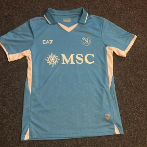 Blå fotbollströja från EA7 - Snygg blå fotbollströja från EA7 med vita detaljer och MSC-logga på framsidan. Tröjan har en klassisk krage och korta ärmar med vita inslag. 