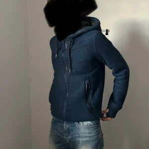 Blå stickad hoodie från Superdry - Säljer en snygg stickad & fodrad hoodie från Superdry med dragkedja och huva. Jackan har patch på ärmen och broderad logga på ryggen. Perfekt för kyliga dagar! 