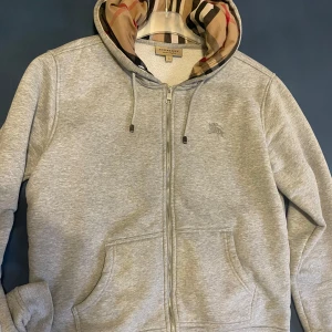 Grå hoodie med dragkedja från Burberry - Snygg grå hoodie från Burberry med dragkedja och klassiskt rutigt mönster i huvan. Perfekt för en stilren och bekväm look. Den har snören i huvan och två fickor framtill.