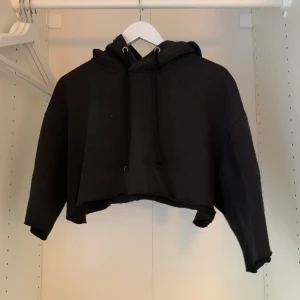 Svart croppad hoodie med tre kvarts ärmar - Säljer en svart croppad hoodie med huva och dragsnören. Perfekt för en avslappnad stil. Den har långa ärmar och en lös passform som ger en bekväm känsla. Passar bra till jeans eller leggings för en casual look.