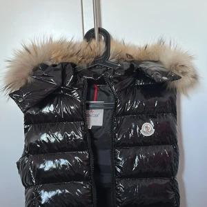 Svart dunväst med pälsdetalj från Moncler - Snygg svart dunväst från Moncler med glansig yta och pälsdetalj på huvan. Perfekt för kyliga dagar när du vill hålla stilen. Västen har en dragkedja framtill och är idealisk för både höst och vinter.