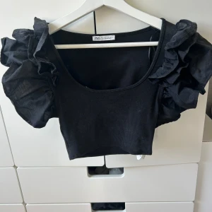Svart croppad topp från Zara - Snygg svart croppad topp från Zara med puffiga volangärmar. Toppen har en rund halsringning och är tillverkad i ett mjukt material.