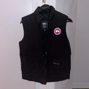 Svart väst från Canada Goose - En svart dunväst från Canada Goose med ett klassiskt märke på bröstet. Västen har en hög krage och stängs med dragkedja och knappar. Perfekt för kyliga dagar. Västen är i alldeles utmärkt skick, inga synliga defekter. Vänliga kom DM om ni vill se fler bilder eller om ni har några frågor. Priset kan diskuteras. Tar gärna emot byten.