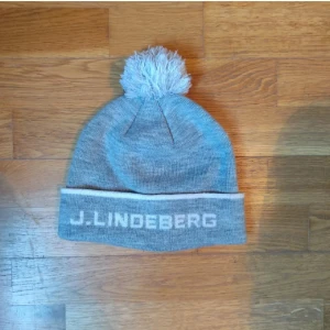 Grå mössa från J.Lindeberg - Snygg grå mössa från J.Lindeberg med en fluffig tofs på toppen. Mössan har en uppvikt kant med broderad logotyp i vitt. Perfekt för en stilren look lätt att styla till många outfits. Helt som ny nästan aldrig använd, skulle säga att jag använt den typ 3 gånger bara säljer den för jag inte riktigt gillar att ha mössa personligen.