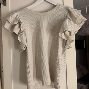 Beige ribbad topp från Zara - Säljer en stilren beige ribbad topp från Zara med volangdetaljer på ärmarna. Perfekt för en chic och avslappnad look. Toppen har en rund halsringning och korta ärmar med volanger.