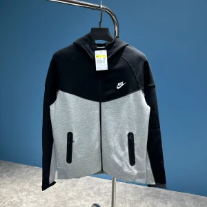 Grå och svart huvtröja från Nike - Snygg huvtröja från Nike i grått och svart med dragkedja framtill. Tröjan har en klassisk design med Nike-logga på bröstet och praktiska fickor med dragkedja. Perfekt för en sportig look. Finns alla storlekar. S M L Xl XLL