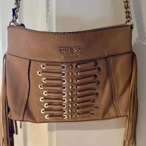 Beige axelväska från Guess - Snygg beige axelväska från Guess med fransar och dekorativa snördetaljer på framsidan. Väskan har en justerbar axelrem med kedjedetaljer och dragkedja upptill. Perfekt för att ge din outfit en trendig touch.