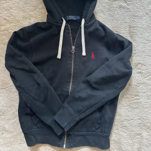 Svart hoodie från Polo Ralph Lauren - Säljer en svart hoodie från Polo Ralph Lauren med dragkedja och vit snörning. Den har en röd broderad logotyp på bröstet och praktiska fickor framtill. Perfekt för en avslappnad stil.