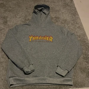 En snygg grå hoodie från, Thrasher, skate - Säljer en grå hoodie från Thrasher med den ikoniska loggan i gult och rött på framsidan. Perfekt för en avslappnad stil med en skön passform och huva. Passar perfekt för skate eller chill dagar. Tröjan har inga snören. Jag skulle uppskatta att tröjan är i M. Hör av dig vid intresse! 🔥och priset är såklart inte hugget i sten