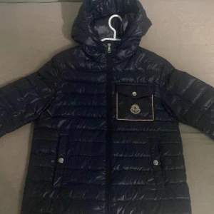 Svart dunjacka från Moncler - Snygg marinblå dunjacka från Moncler med dragkedja och tryckknappar. Jackan har en praktisk huva och en stilren ficka med Moncler-logga på bröstet. Perfekt för kyliga dagar. Kan sänka pris om det behövs.