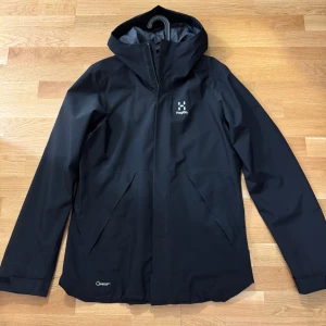 Svart jacka från Haglöfs - Säljer en stilren svart regn/vindjacka från Haglöfs. Jackan har en praktisk huva och dragkedja framtill. Perfekt för regniga dagar med sin vattentäta design. Den har en enkel och funktionell stil med diskreta fickor. Storlek S/Nypris 2899kr