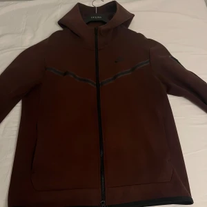 Brun/vinröd nike tech - Säljer en brun/vinröd nike tech med dragkedja och långa ärmar. Den har en stilren design med diskreta detaljer och Nike-logga på bröstet. Perfekt för en sportig look.