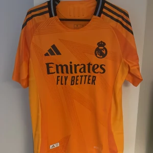Real Madrid tröja 24/25 - Säljer en orange Real Madrid fotbollströja 24/25 från Adidas med Vini Jr. Heläkta och i nyskick. Tröjan är spelarversion, alltså den spelarna har på sig och därför den dyrare versionen. Stolek M.