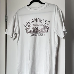 Vit t-shirt med Los Angeles-tryck - Säljer en vit t-shirt med tryck av 'Los Angeles California' och 'Venice Beach 86'. T-shirten har korta ärmar och ett avslappnat tryck med palmer och hus. Perfekt för en casual look. Väldigt oversized och mysig. Storlek: XS. Axel: 51cm. Längd: 75cm.