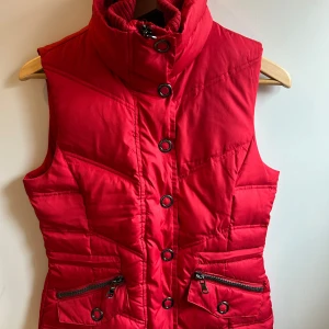 Röd quiltad väst från Esprit - Snygg röd quiltad väst från Esprit med hög krage och dragkedja. Västen har knappar och praktiska fickor med dragkedjor. 
