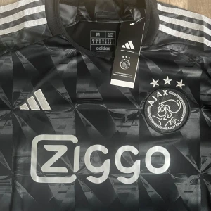 Svart Ajax fotbollströja från Adidas - Snygg svart Ajax fotbollströja från Adidas med korta ärmar och ett geometriskt mönster. Tröjan har vita detaljer och Ziggo-logga på framsidan. Perfekt för fotbollsfans som vill visa sitt stöd för laget.