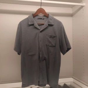 Grå kortärmad skjorta från 157 - Säljer en stilren grå kortärmad skjorta från 157. Skjortan har en klassisk krage och knappar framtill. Den är utrustad med en bröstficka för extra stilpoäng. Perfekt för en avslappnad look.