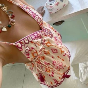 Mönstrad ärmlös topp - Säljer en färgglad ärmlös topp med broderade detaljer i rosa och orange på en beige bas. Toppen har smala axelband och en djup V-ringning med dekorativa kanter. Perfekt för varma sommardagar! storleken är oklar, men skulle säga M. Skriv privat för egna bilder! 