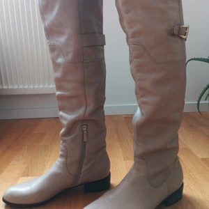 Beige stövlar med spänne läder  - Snygga beige stövlar i skinn med dragkedja och dekorativt spänne upptill. Perfekta för höst och vinter med en stilren design och låg klack.