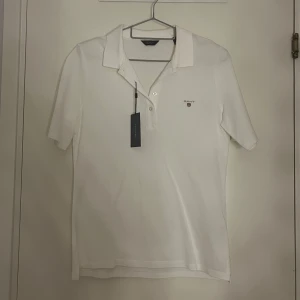Vit pikétröja från Gant - Säljer en klassisk vit pikétröja från Gant med korta ärmar och en liten logotyp på bröstet. Tröjan har en stilren design med krage och knappar framtill. Perfekt för en avslappnad men stilfull look.
