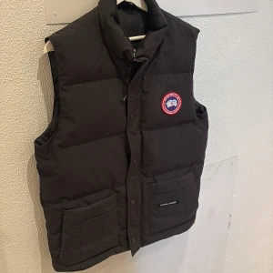 Svart dunväst från Canada Goose - Säljer en svart dunväst från Canada Goose med deras ikoniska logga på bröstet. Västen har en dragkedja och knappar framtill samt två praktiska fickor. Perfekt för kyliga dagar.