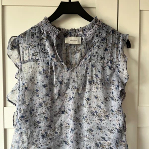 Blommig blus från Neo Noir - Säljer en söt blommig blus från Neo Noir i ljusblått med små blommor i mörkare nyanser. Blusen har en v-ringad design med volangdetaljer vid halsen och ärmlös stil. Perfekt för en sommaren!💞💞