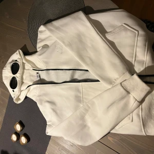 Vit hoodie med dragkedja från C.P. Company - Säljer en snygg vit hoodie från C.P. Company i mycket bra skick. Den har en cool dragkedja och ikoniska glasögon i huvan. Perfekt för höst och vår, med en normal passform och långa ärmar. Grym för både chill och casual outfits!