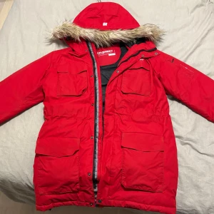 Röd parkajacka från Everest - Säljer en snygg och varm röd parkajacka från Everest i storlek 36. Jackan har en mysig fuskpäls på huvan och flera praktiska fickor. Perfekt för kalla vinterdagar! Den är i mycket bra skick och har en dragkedja framtill. Stygnen vid höger ficka är lite slitna men det är inget som påverkar utseende eller jackans funktion.
