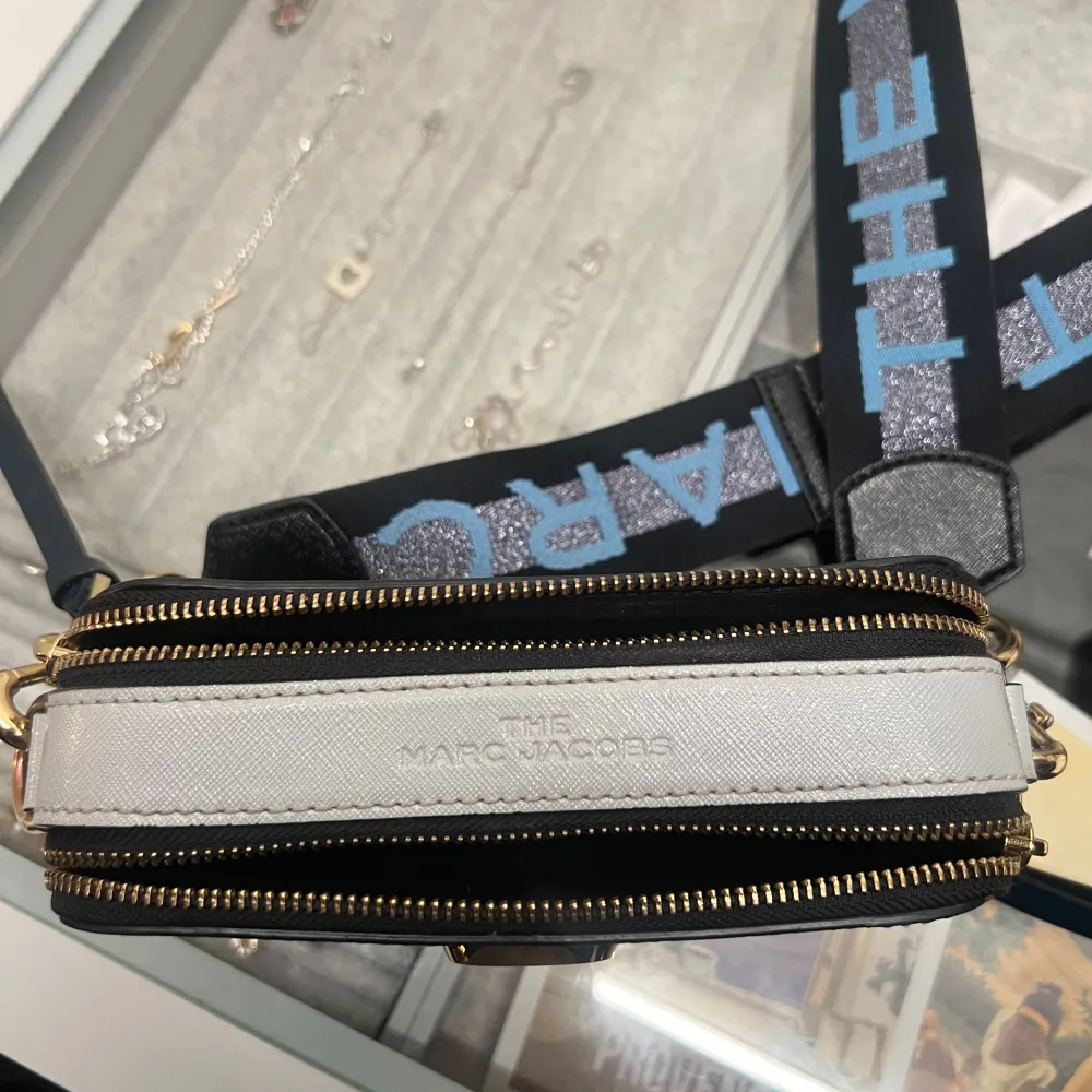 Snygg marinblå axelväska från Marc Jacobs med ett stort guldspänne framtill. Väskan har en bred, justerbar axelrem med text i blått och silver. Den är perfekt för både vardag och fest. Två dragkedjor upptill ger enkel åtkomst till dina saker.. Laukut & Käsilaukut.