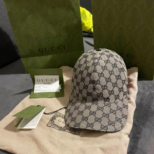 Gucci keps i bästa skick. - Säljer en snygg Gucci keps i mycket bra skick. Den har det klassiska GG-monogrammet i beige och svart, med en röd och grön rand på sidan. Perfekt för att ge din outfit en lyxig touch. Kepsen kommer med originalförpackning och etiketter.