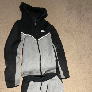 Nike tech fleece ungdom - Säljer en snygg och bekväm grå och svart hoodie från Nike. Den har en dragkedja framtill och en klassisk Nike-logga på bröstet. Perfekt för träning eller en avslappnad dag. Hoodien har långa ärmar och en justerbar huva. Passar perfekt till höst och vår!