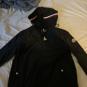 Svart vindjacka från Moncler - Säljer en snygg svart vindjacka från Moncler. Jackan har en dragkedja framtill och en huva med röd och vit rand. Den är i mycket bra skick och perfekt för höst och vår. Moncler-loggan finns på ärmen och insidan. Passar perfekt för en stilren look!