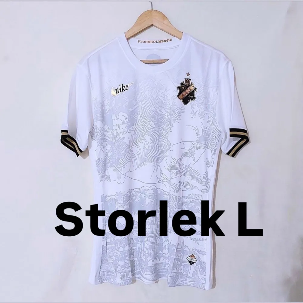 Säljer en snygg vit fotbollströja från Nike i storlek L. Tröjan har ett unikt mönster i grått och detaljer i guld på bröstet. Ärmarna är korta med svarta och guldiga ränder. Perfekt för både träning och vardag!. T-shirts.