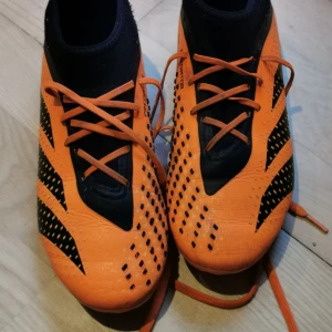 Orange fotbollsskor med svarta detaljer - Säljer ett par grymma fotbollsskor i orange med svarta detaljer. De har snörning och en cool design med svarta mönster. Skorna har dobbar för bra grepp på planen. Perfekta för fotbollsträning eller match. Tillverkade i syntetmaterial för hållbarhet och lätt vikt.