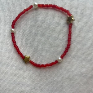 Rött pärlarmband med stjärndetaljer - Ett sött armband med små röda pärlor och vita pärlor, dekorerat med guldstjärnor. Perfekt för att lägga till en färgglad touch till din outfit. Passar bra till både vardag och fest!
