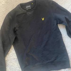 Svart tröja från Lyle & Scott - Säljer en svart tröja från Lyle & Scott i bra skick. Den har en gul logga på bröstet och är perfekt för både vardag och fest. Tröjan är bekväm och stilren, ett måste i garderoben! Str xs