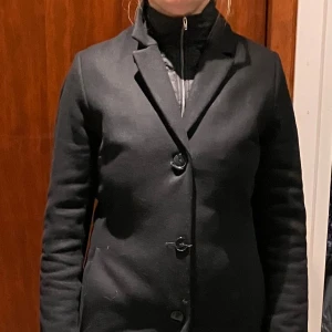 Svart kappa med knappar - Säljer en stilren svart kappa i ull i mycket bra skick. Det finns en krage att sätta fast för blåsiga dagar. Varm och skön och kan användas höst, vinter och vår. 