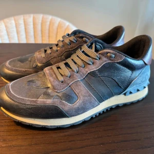 Valentino Rockrunner i mocka och skinn - Säljer ett par snygga Valentino Rockrunner sneakers i en mix av mocka och skinn. Skorna har en cool kombination av svart och grått med en touch av brunt på hälen. De har snörning och en bekväm sula som passar perfekt för både vardag och fest. Perfekt för dig som vill ha stil och komfort i ett!