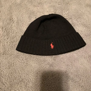 Svart mössa från Ralph Lauren - Snygg svart mössa från Ralph Lauren med röd broderad logga. Perfekt för kyliga dagar, den är både stilren och varm. Passar till alla outfits och är ett måste i vintergarderoben!