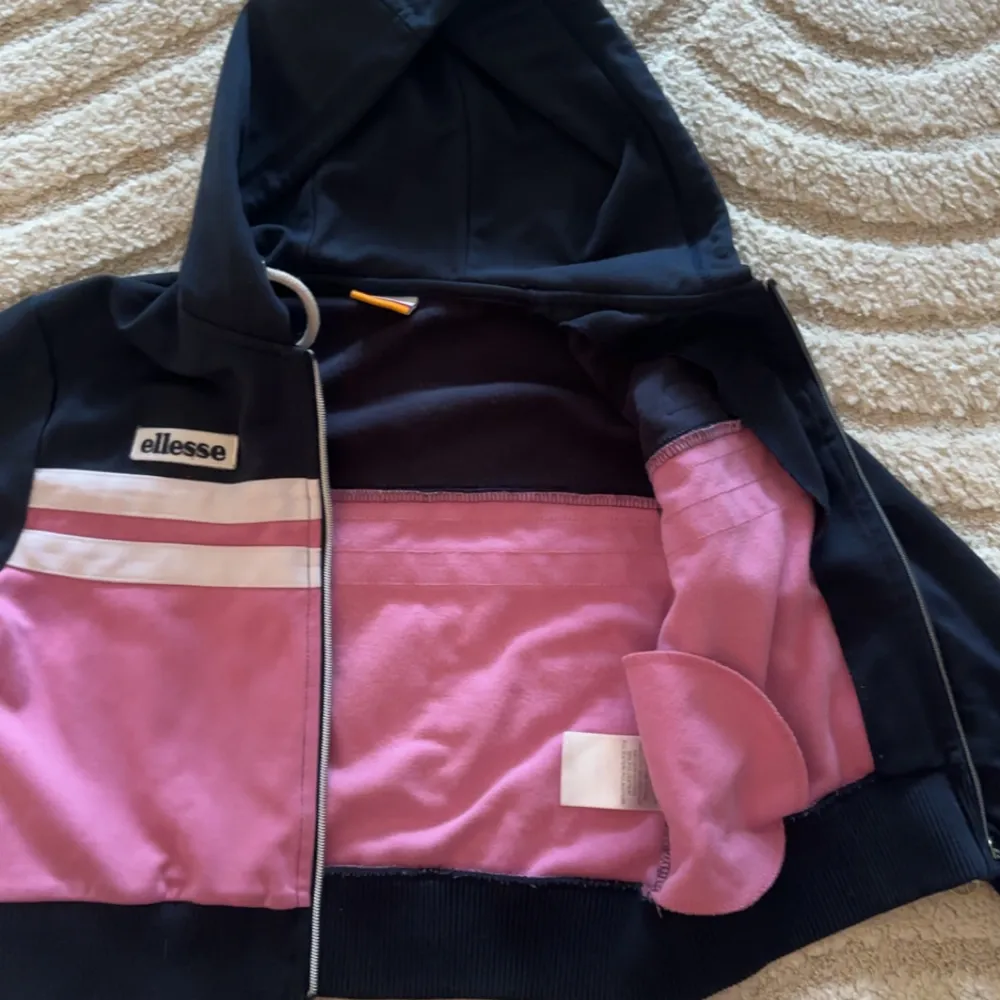 Säljer en snygg och bekväm hoodie från Ellesse i svart och rosa. Den har en dragkedja framtill och en justerbar huva med vita snören. Perfekt för en avslappnad stil eller träning. Den är croppad och har ribbade muddar vid ärmslut och nederkant💗. Tröjor & Koftor.