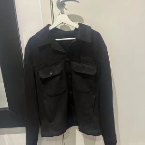 Svart overshirt med knappar - Säljer en stilren svart overshirt med knappar framtill. Den har två stora bröstfickor med lock och knappar, vilket ger en cool och praktisk look. Perfekt för höst och vår, och funkar både till vardags och lite mer uppklädda tillfällen. Passar bra över en hoodie eller t-shirt. Från zara. Endast testad men fel storlek. Storlek L
