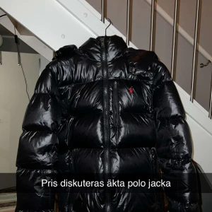 Svart dunjacka från Polo Ralph Lauren - Säljer en äkta svart dunjacka från Polo Ralph Lauren i storlek M. Jackan är i mycket bra skick och har en glansig finish med en röd logga på bröstet. Den har en dragkedja framtill och är perfekt för kalla vinterdagar. Hör av dig för prisdiskussion!