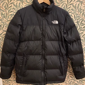 Svart dunjacka från The North Face - Säljer en svart dunjacka från The North Face i mycket bra skick. Jackan har en dragkedja framtill och fickor med dragkedja. klassisk pufferstil medium tjocklek som håller dig varm och bekväm.  Yttertyg i nylon, dun padding i kanaler som gör den lätt men gosig. Perfekt för både vardag och utomhusaktiviteter!