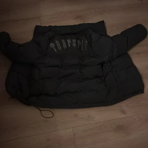 Svart pufferjacka från Tripstar - Säljer en svart pufferjacka från Tripstar i bra skick. Jackan har en avtagbar huva och dragkedja framtill. Perfekt för kalla dagar med sin tjocka vaddering. Den har också en snygg broderad logga på ryggen. Passar både till vardags och lite mer avslappnade tillfällen.
